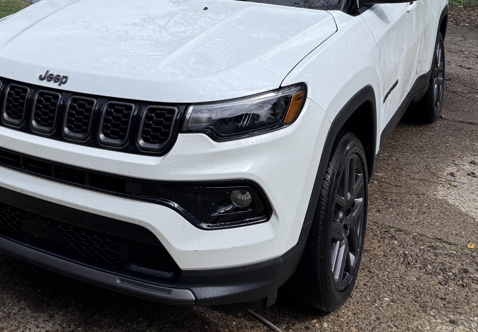 Front of a clean white Jeep Compass, close on the grille and Jeep badge, with trees reflected in the hood.