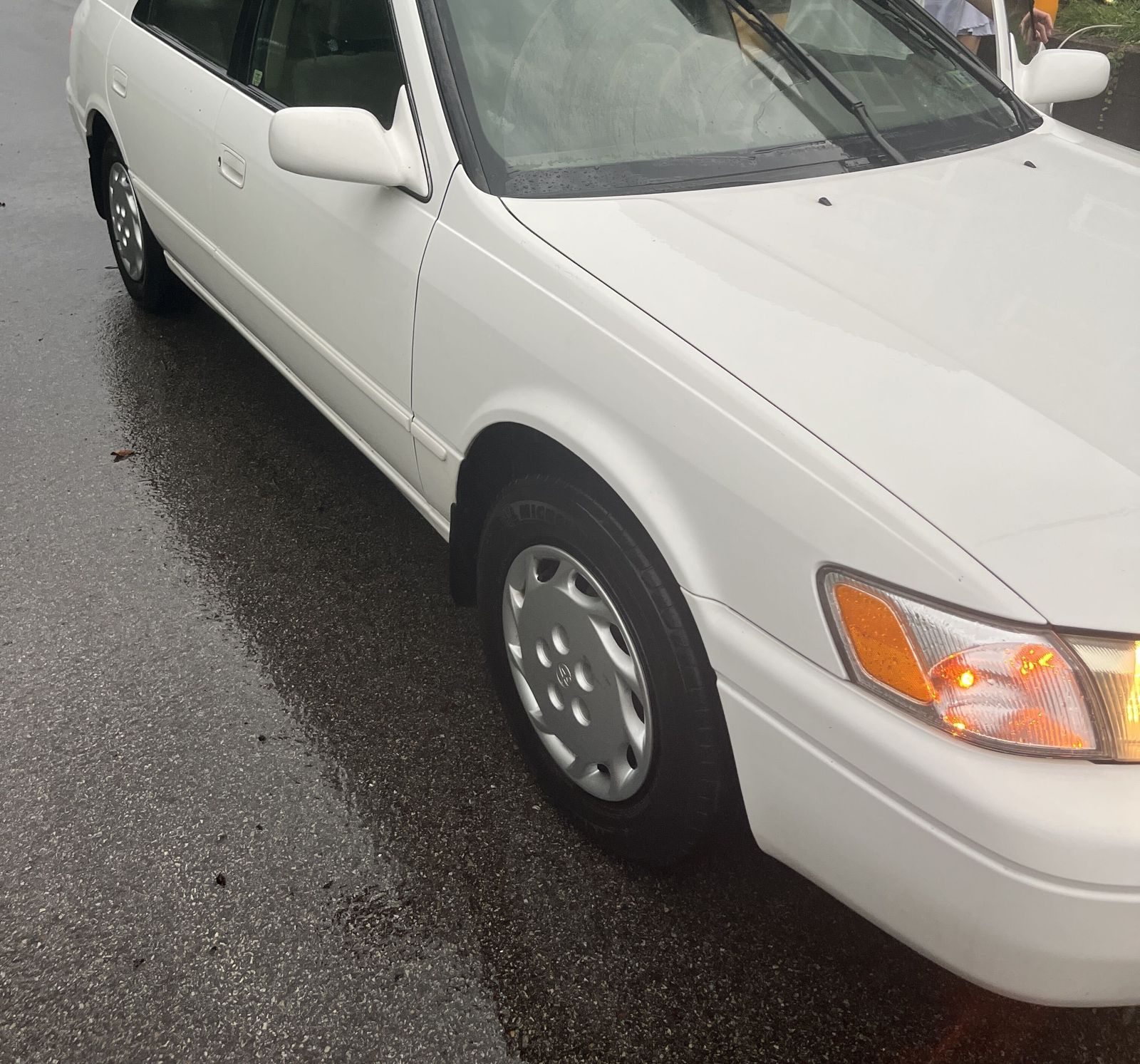 White Toyota Camry after the wash, front three quarter view on a wet road.