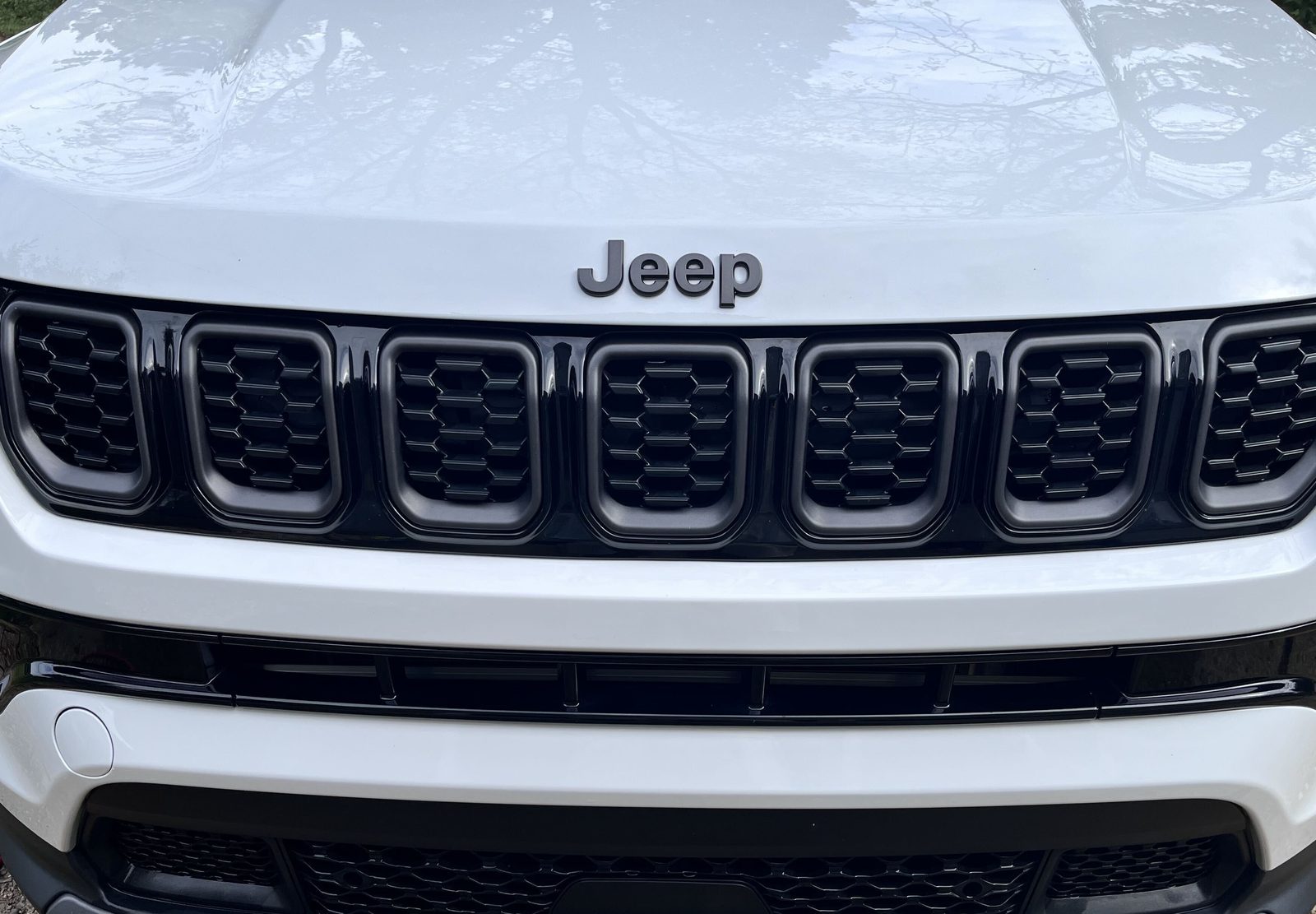 Front of a clean white Jeep Compass, close on the grille and Jeep badge, with trees reflected in the hood.