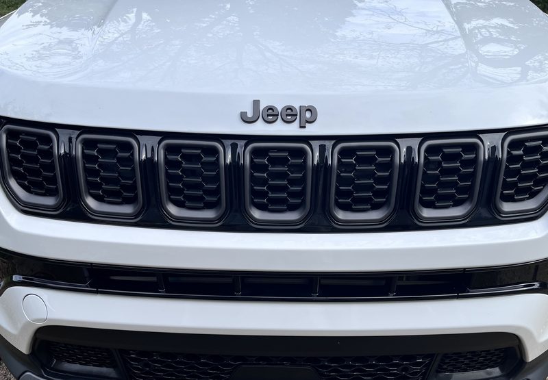 Front of a clean white Jeep Compass, close on the grille and Jeep badge, with trees reflected in the hood.